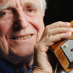 Foto av Douglas Engelbart som gammal. Han håller i den första musen, som delvis är av trä.